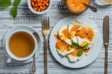 Slices of adyghe cheese poured over sea buckthorn jam with green tea in white cup and fresh raw sea buckthorn berries during breakfast on wooden table, close-up, top view. Healthy food concept