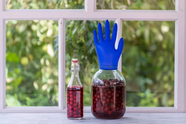 Homemade tincture of red cherry in a jar with a glove shaped shutter. Berry alcoholic drinks concept. Homemade red wine made from ripe cherries in glass jar on white wooden window background, Ukraine