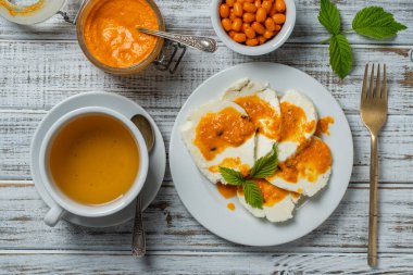 Slices of adyghe cheese poured over sea buckthorn jam with green tea in white cup and fresh raw sea buckthorn berries during breakfast on wooden table, close-up, top view. Healthy food concept
