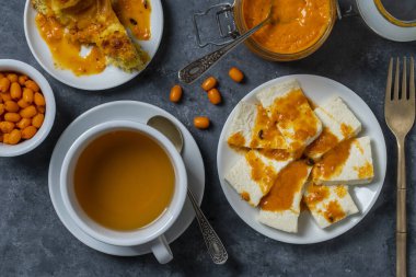 Slices of adyghe cheese poured over sea buckthorn jam with green tea in white cup and fresh raw sea buckthorn berries during breakfast on table, close-up, top view. Healthy food concept