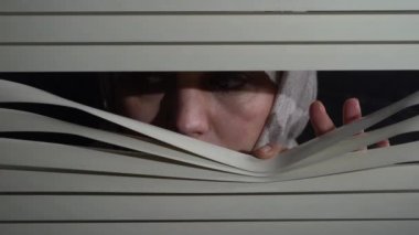 Kapalı alandaki başörtülü esmer kadının ön portresini kapat geceleri elleriyle pencereye panjur iterek. Dışarıdan korkunç görünüyor.