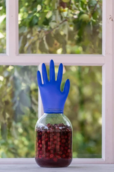 Homemade tincture of red cherry in a jar with a glove shaped shutter. Berry alcoholic drinks concept. Homemade red wine made from ripe cherries in glass jar on white wooden window background, Ukraine