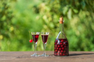 İki bardakta ev yapımı vişneli brendi ve yaz bahçesindeki ahşap bir masada cam bir şişede.