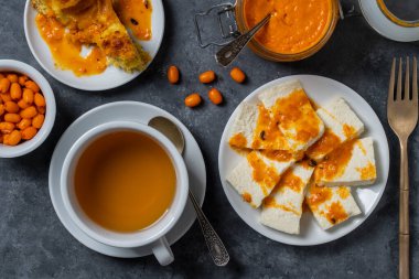 Slices of adyghe cheese poured over sea buckthorn jam with green tea in white cup and fresh raw sea buckthorn berries during breakfast on table, close-up, top view. Healthy food concept