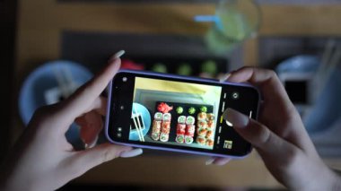 Kız, rahat Japon restoranında akıllı telefon kullanarak suşi fotoğrafı çekiyor. Kadın eli, modern akıllı telefonla yiyeceklerin fotoğrafını çeker. Yaklaş, 4K