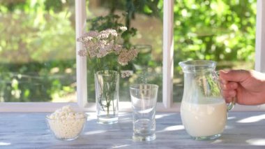 Pouring fresh milk into a transparent glass from jug on the windowsill by the window of the house on a summer day near the garden, close up