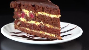 Delicious dark chocolate cream cake from short pastry with custard and cherry berry in the home kitchen. Piece of dark chocolate cake on the black background rotating. Homemade baking. Close up