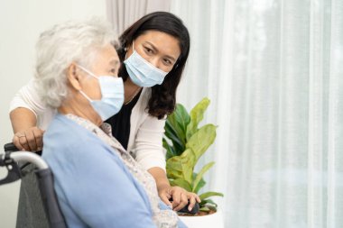 Bakıcı, tekerlekli sandalyede oturan ve güvenlik enfeksiyonu Covid-19 Coronavirus 'u korumak için maske takan yaşlı ve yaşlı bir kadına yardım ediyor..
