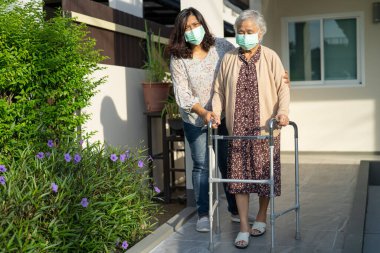 Bakıcı, Asyalı yaşlı ya da yaşlı bir kadına rampada yürüyen bir kadınla yürümesinde ve güvenlik enfeksiyonu Covid-19 Coronavirus 'u korumak için maske takmasında yardım ediyor..