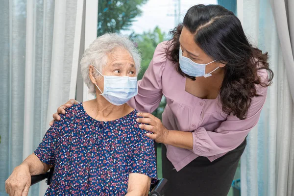 Bakıcı, Asyalı yaşlı ya da yaşlı kadın tekerlekli sandalyede oturan ve güvenlik enfeksiyonunu korumak için maske takan Covid19 Coronavirus 'a yardım ediyor..