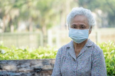 Asyalı yaşlı bir kadın bankta oturuyor ve güvenlik enfeksiyonunu korumak için maske takıyor Covid-19 Coronavirus parkta sosyal mesafeyi koruyor..