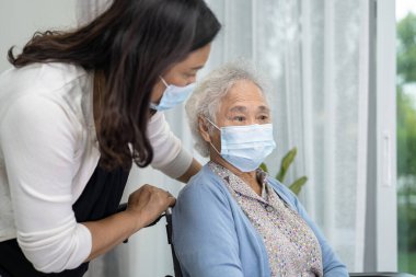 Tekerlekli sandalyede oturan ve güvenlik enfeksiyonunu korumak için maske takan yaşlı ve yaşlı bir kadına yardım edin..