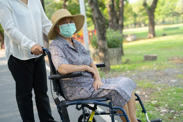 Tekerlekli sandalyedeki yaşlı ve yaşlı kadına yardım edin ve güvenlik enfeksiyonunu korumak için maske takın. Parkta Covid19 Coronavirus.