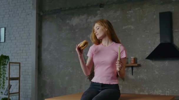 ビデオだ。キッチンに座っている女性は、ハンバーガーや健康的な食べ物を選ぶべきか分からないジレンマを抱えています。赤い髪だ。健康的なライフスタイル — ストック動画