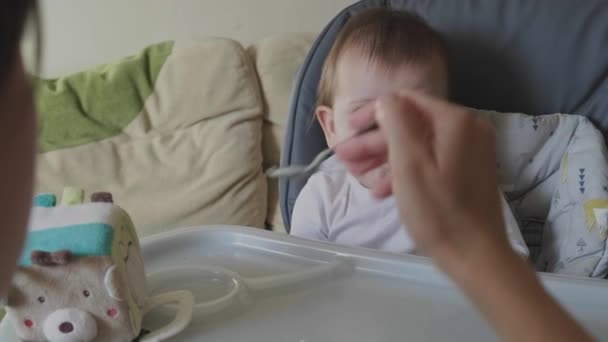 Vídeo. Mães mãos alimentando o bebê com purê de frutas sentado na mesa especial para crianças e olhando cuidadosamente para a câmera. Miúda. Refeição saudável. — Vídeo de Stock