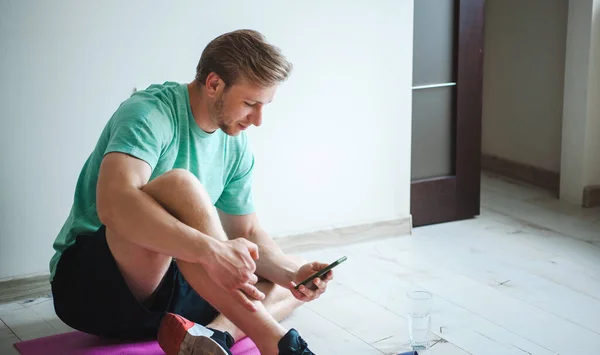 Uomo rilassante dopo aver fatto allenamento online al mattino seduto sul pavimento e guardando il telefono con un bicchiere d'acqua — Foto Stock