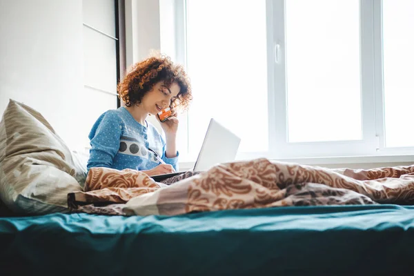 Mujer rizada hablando por teléfono directamente desde la cama notando la tarea. Clases en línea — Foto de Stock