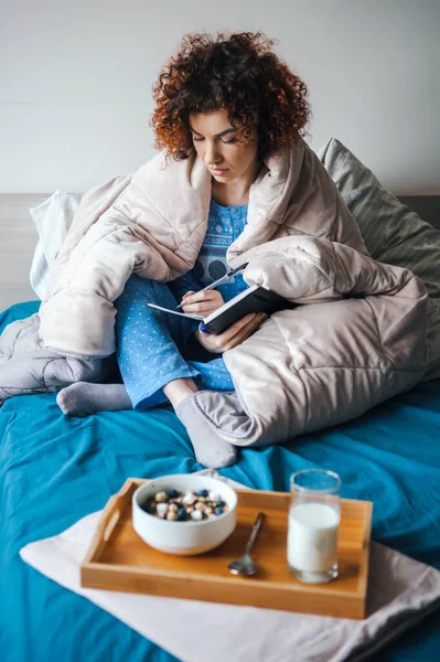 Wanita keriting yang menghabiskan akhir pekan pagi di tempat tidur di bawah selimut menulis dalam buku hariannya. Sarapan di tempat tidur. Sarapan sehat — Stok Foto