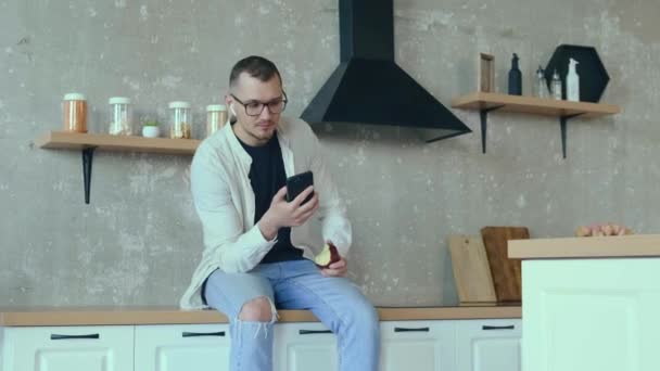 Vídeo. Um homem a passar a pausa para o almoço na cozinha a comer uma maçã e a escrever mensagens ao telefone. Ligação Bluetooth. Tecnologia da Internet. — Vídeo de Stock