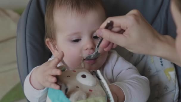 Video con un bebé desayunando directamente de la cucharadita estirada en su boca por las manos de sus madres. Cuidado del bebé. Madre hija amor. Divertido bebé feliz. — Vídeos de Stock