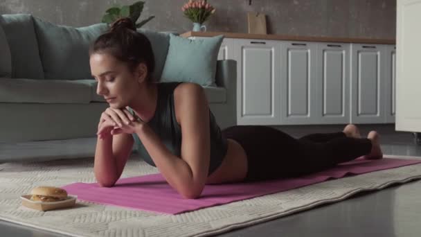 Video van een vrouw liggend op de fitness matras met haar handen onder haar kin kijkend naar de hamburger voor haar. Gezonde levensstijl — Stockvideo