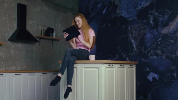 Відео з жінкою, яка проводить вільний час на кухні, де п'є каву і робить покупки в Інтернеті на планшеті. Сучасний жіночий спосіб життя. Сучасна кухня — стокове відео