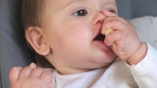 Video con un bebé tumbado en la cama mirando con grandes ojos negros directamente a la cámara llevando su mano a la boca comiendo plátano. Cara de bebé — Vídeos de Stock