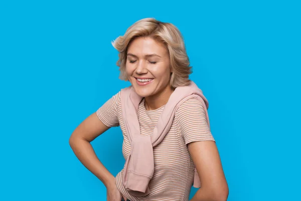 Mujer con una camiseta a rayas aislada sobre un fondo azul, sonriendo con los ojos cerrados y sosteniendo las manos sobre las caderas. Cara de belleza — Foto de Stock