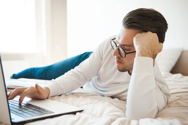 Hombre con gafas acostado en la cama en el portátil navegando por Internet. Tecnología de negocios. Comunicación inalámbrica. — Foto de Stock