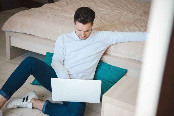 Hombre sentado en el suelo junto a la cama trabajando en el ordenador. Ambiente de trabajo en casa. Comunicación en línea —  Fotos de Stock
