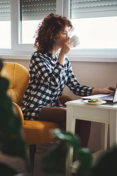 Вид сбоку на молодую женщину, отдыхающую на работе, чтобы выпить чашечку кофе и съесть сэндвич. Интернет-технологии. — стоковое фото