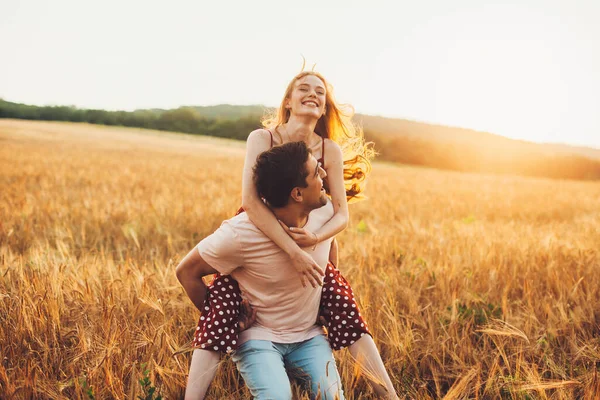 Νεαρό ζευγάρι διασκεδάζει στο χωράφι με τα σιτηρά. Βόλτες με το γουρουνάκι. Οικογενειακό Σαββατοκύριακο. — Φωτογραφία Αρχείου
