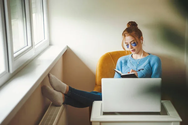 Framifrån på en ung kvinna som studerar hemifrån vid datorn, sitter på fåtöljen och antecknar och lyssnar på musik — Stockfoto