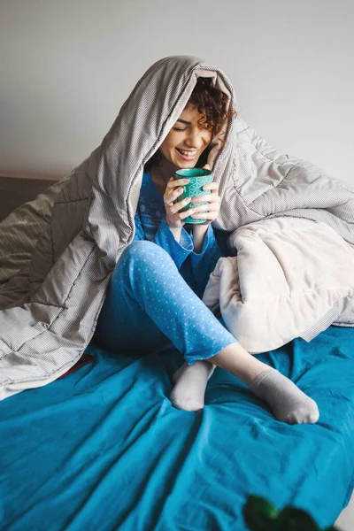 Mañana de una mujer rizada cubierta por la manta disfrutando de una bebida caliente — Foto de Stock