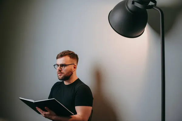 Hombre de negocios caucásico con barba en camiseta negra sentado en silla contra el fondo de una pared blanca iluminada por una lámpara. Vista lateral. Usar —  Fotos de Stock