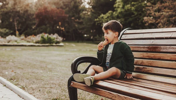 Vista lateral de um menino caucasiano comendo sorvete sentar no banco de madeira no parque. Recreação ao ar livre. — Fotografia de Stock