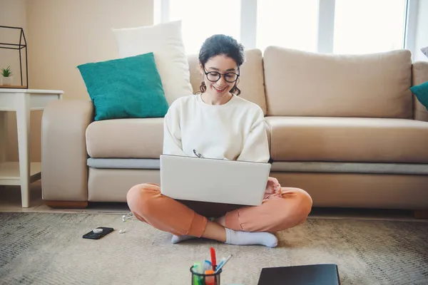Вид спереди женщины, делающей заметки, опираясь на диван и сидя на полу с ноутбуком на скрещенных ногах. Онлайн-образование. Дистанционное образование. — стоковое фото