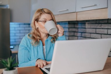 Orta yaşlı bir kadın evde laptopuyla telgraf çekerken kahve içer. İş kadını portresi. Evden çalışmak