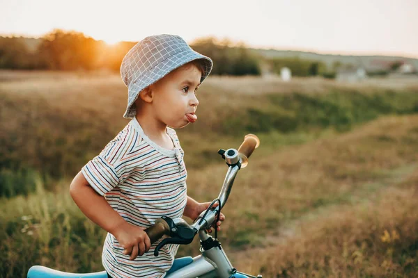 Bir köy yolunda bisiklet süren mavi şapkalı ve dilini gösteren bir çocuğun portresi. Çocukluk — Stok fotoğraf