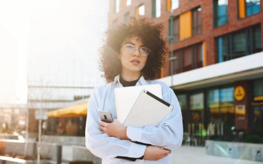İşten eve elinde dizüstü bilgisayar ve telefonla dönen kıvırcık bir kadının ön görüntüsü. Gün batımı ışığı.