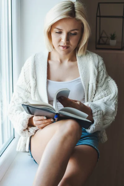 Mujer rubia blanca sentada en el alféizar de la ventana y leyendo un libro. Autoaislamiento. Coronavirus. Vista frontal. — Foto de Stock