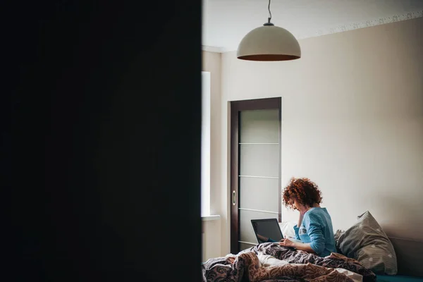 Vista posterior de la mujer caucásica usando el ordenador portátil sentado en la cama en casa. Mañana. Enterradora. Vista desde la puerta medio abierta — Foto de Stock