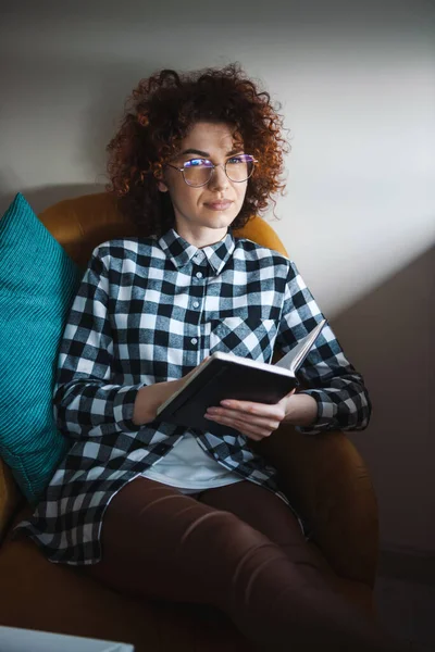 Lockig kvinna med glasögon sittandes i fåtöljen hemma och skriver anteckningar. Coronavirus. Coronavirus pandemi. E-lärande — Stockfoto
