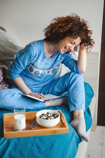 Potret wanita muda tersenyum duduk di piyama biru di tempat tidur, membaca buku yang menarik. Sarapan di tempat tidur. Waktunya tidur. Rambut keriting. — Stok Foto