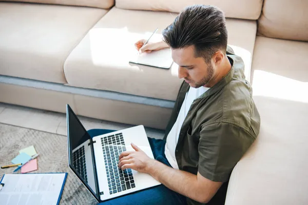 Sidovy av en man som deltar i en online-konferens, anteckningar, sitter på golvet lutande på soffan. Utbildning på nätet. Koncept för hemmakontor — Stockfoto