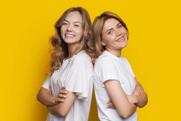 Porträt zweier lachender Frauen in weißen T-Shirts, die Rücken an Rücken vor gelbem Hintergrund stehen und die Arme verschränkt halten. Nahaufnahme. — Stockfoto