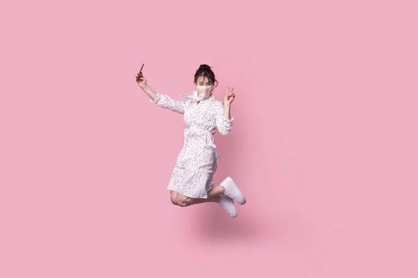 Фотографія леді стрибає під час фотографії з телефоном, показує v-знак, дивлячись на камеру носить медичну маску, плаття, кросівки, ізольовані на рожевому — стокове фото