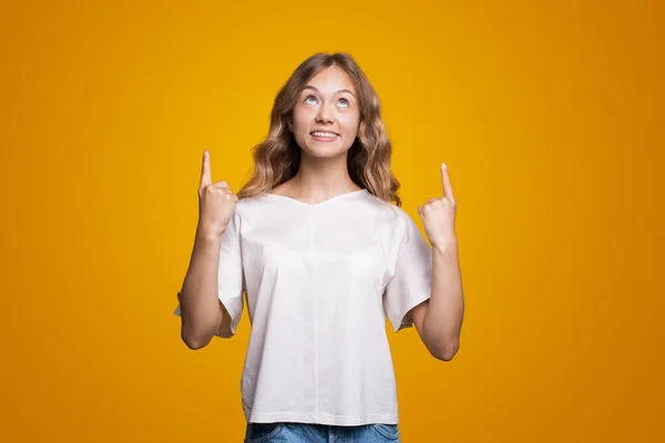 Kaukasische Frau im weißen T-Shirt blickt nach oben und zeigt mit dem Zeigefinger auf den Kopierraum vor gelbem Hintergrund. Gestik und Mimik — Stockfoto