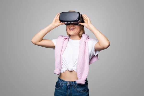 Mujer caucásica sonriente que obtiene experiencia usando gafas de realidad virtual con auriculares VR sobre fondo gris. Juego, realidad virtual. — Foto de Stock