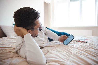 Yatakta yatan ve ona telefonda gönderilen bazı belgeleri inceleyen bir adam. İş teknolojisi. Evden çalış.